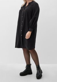 Femme portant une robe noire à boutons, arrivant au genou, des collants noirs transparents et des bottines à plateforme noires avec des zips à l'avant.