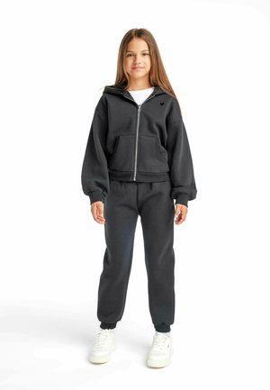 Veste à capuche noire zippée avec poches avant et joggers assortis, présentant une coupe décontractée et des chevilles resserrées, le tout associé à des baskets blanches.
