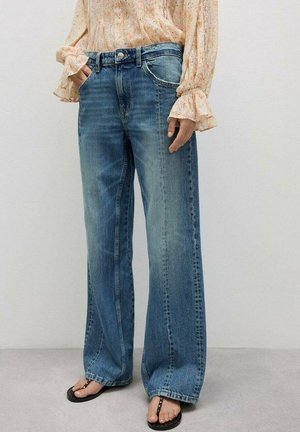 Niebieskie dzwony z szerokimi nogawkami noszone z kwiecistą bluzką z długim rękawem i czarnymi sandałami z ćwiekami na szarej podłodze i gładkim tle ściany.