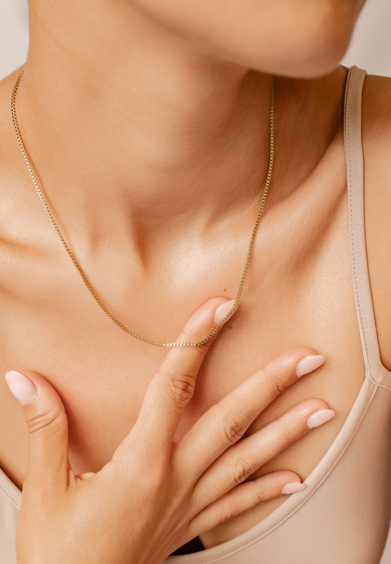 Goldene Kettenhalskette mit einem zarten, flachen Design, die auf der Haut ruht. Glatte Oberfläche und sanfter Glanz; Finger berühren die Kette.