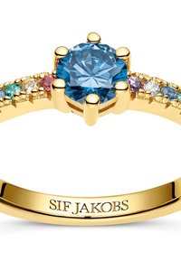 Anello in oro con una pietra preziosa blu rotonda centrale, affiancata da pietre multicolori più piccole. La scritta incisa all'interno della fascia è "SIF JAKOBS."