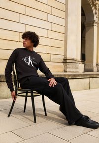 Svarte Calvin Klein Jeans hettegenser med hvit logo, kombinert med løse svarte bukser og svarte sko, sittende på en trestol.