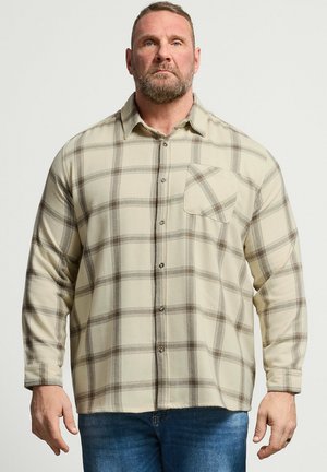 Skæggemand iført beige ternet knap-op-skjorte og blå jeans, stående mod en ensfarvet lys baggrund.