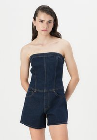 Salopette in denim con design senza spalline, di colore blu scuro, corpino aderente, gambe corte e cuciture a contrasto. Senza tasche visibili.