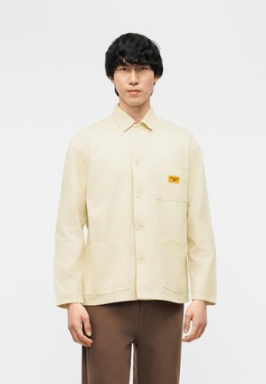 Jaunas vyras dėvi kremiško spalvos užsegamą striukę su trimis priekinėmis kišenėmis ir maža geltona etikete ant krūtinės, derina su rudomis kelnėmis.