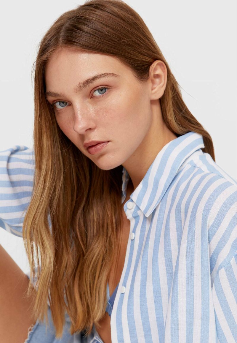 Biała i niebieska koszula w paski z luźnym krojem, z kołnierzykiem i podwiniętymi rękawami, zestawiona z niebieską bluzką pod nią.
