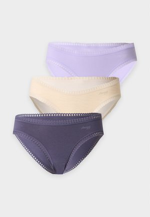 Femme debout pieds nus portant un bralette turquoise et une culotte gris foncé ornée de dentelle sur un fond clair uni.