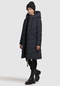 Manteau long rembourré en marine foncé, doté d'une capuche, d'une fermeture éclair à l'avant et d'une poche latérale avec rabat. Porté par-dessus des leggings noirs et des bottes imposantes.