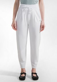 Person iført hvide højtaljede bukser med snøre og sorte balletsko, stående mod en ensfarvet lys baggrund.