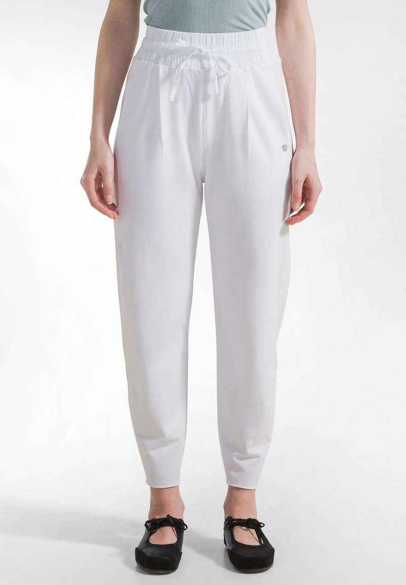 Person iført hvide højtaljede bukser med snøre og sorte balletsko, stående mod en ensfarvet lys baggrund.