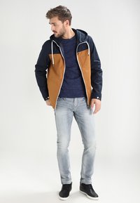 Chaqueta de hombre con bloques de color en azul marino y marrón, con capucha, cremallera frontal y cordones blancos. Lleva puesta una camisa azul marino y jeans claros.