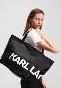 Borsa a tracolla nera con finitura lucida, grande logo bianco "KARL LAGERFELD" e lunghe maniglie nere, tenuta su una spalla.
