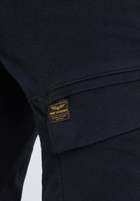 Tamno plavi materijal s teksturiranim finishom, sadrži žutu PME Legend oznaku. Dizajn uključuje džep s preklopom s čistim šavovima.