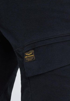 Tissu bleu foncé avec une finition texturée, arborant une étiquette jaune PME Legend. Le design comprend une poche à rabat avec des coutures soignées.