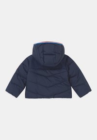 Giacca imbottita blu navy con cappuccio. Presenta cuciture diagonali, un bordo rosso e blu a contrasto lungo il cappuccio e una finitura liscia.