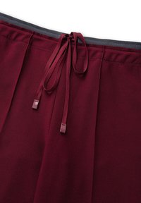 Bordeaux rok met een gladde textuur, elastische tailleband en decoratieve strikken aan de voorkant. Heeft subtiele plooien en een grijze bies.