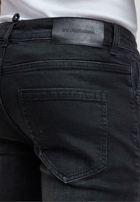 Crne traperice od denima s kožnom oznakom na pojasu, stražnjim džepovima s prošivnim detaljima i uskim krojem.