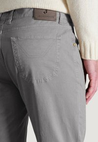 Pantalones de algodón grises con un bolsillo trasero que presenta un diseño de costura distintivo y una etiqueta color miel marcada "JECKERSON", combinados con un suéter crema.