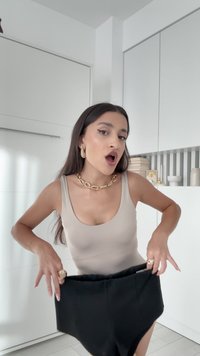 Kobieta z długimi ciemnymi włosami, ubrana w beżowy top na ramiączkach i złotą biżuterię, trzymająca dużą parę czarnych spodni w nowoczesnej białej kuchni.