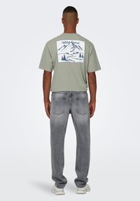 Camiseta de algodón verde claro con un gráfico de montaña en azul marino en la parte posterior, combinada con jeans de mezclilla gris y zapatillas blancas.