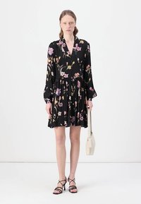 Abito floreale nero con maniche lunghe, scollatura a V e gonna arricciata. Presenta motivi floreali rosa e gialli, abbinato a tacchi neri con cinturini e una borsa bianca.