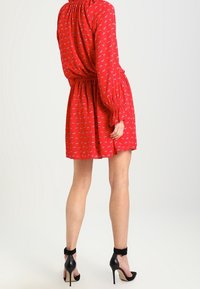 Robe rouge, longueur genoux avec un motif floral, col froncé, taille élastique et manches bouffantes ; associée à des chaussures à talons hauts noires.