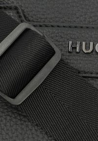 Materiale nero testurizzato con una tracolla intrecciata e accessori metallici che riportano il nome del marchio "HUGO." Si concentra su un design compatto e sulla durata.