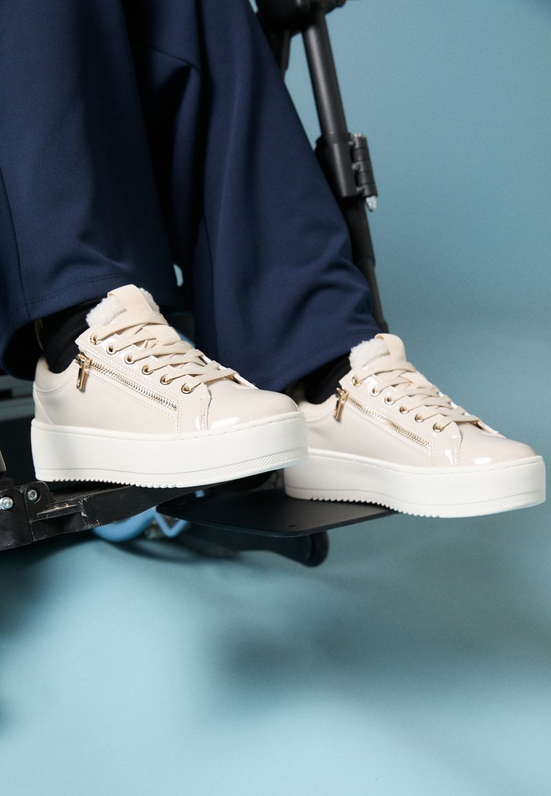 Μπεζ πλατφόρμες sneakers με γυαλιστερή πατίνα, σχέδιο με κορδόνια και λεπτομέρεια με φερμουάρ στο πλάι. Λευκή σόλα με υφή.