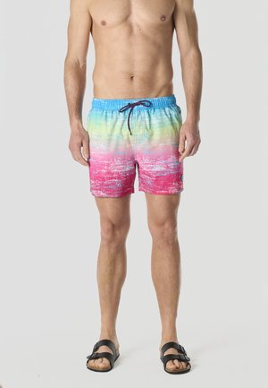 Hombre sin camisa vistiendo pantalones de baño con degradado de colores en tonos azul, verde y rosa, y sandalias negras con hebilla, de pie contra un fondo liso.