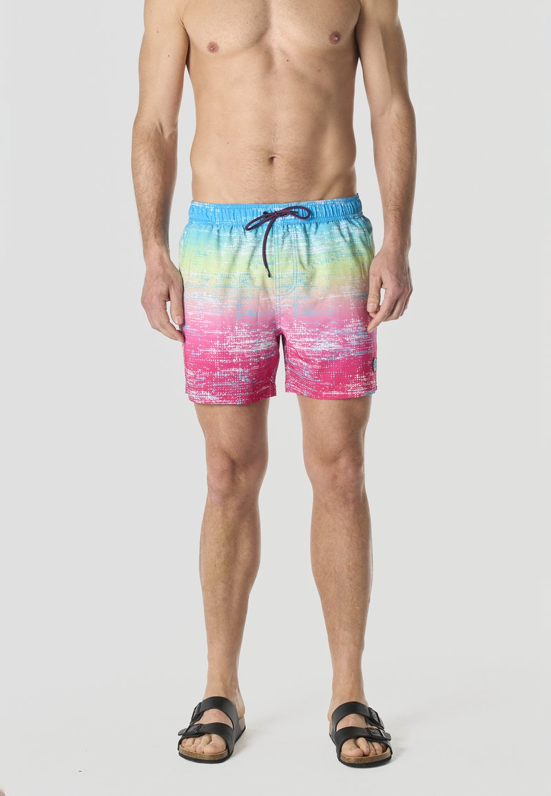 Homme torse nu portant un short de bain à dégradé coloré avec des nuances de bleu, vert et rose et des sandales noires à boucle, debout devant un fond uni.