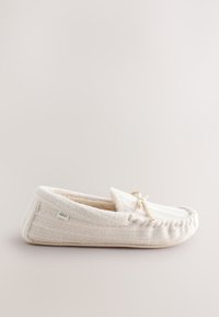 Chausson mocassin doux de couleur crème avec un petit nœud sur le dessus et une étiquette en tissu sur le côté, présenté sur un fond neutre.