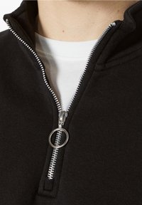 Svart zip-up sweatshirt med hög krage, försedd med en silverdragkedja och cirkulär dragknapp, lager på en vit t-shirt. Mjuk textur synlig.