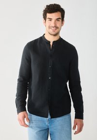 Camisa negra de manga larga con botones y cuello mao, tejido ligeramente texturizado, combinada con jeans de color azul claro. Diseño de ajuste casual.