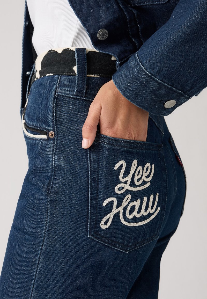 Mørkeblå denimbukser med en hvid "Yee Haw" brodering på baglommen. Har en struktureret talje og kontrasterende syninger.