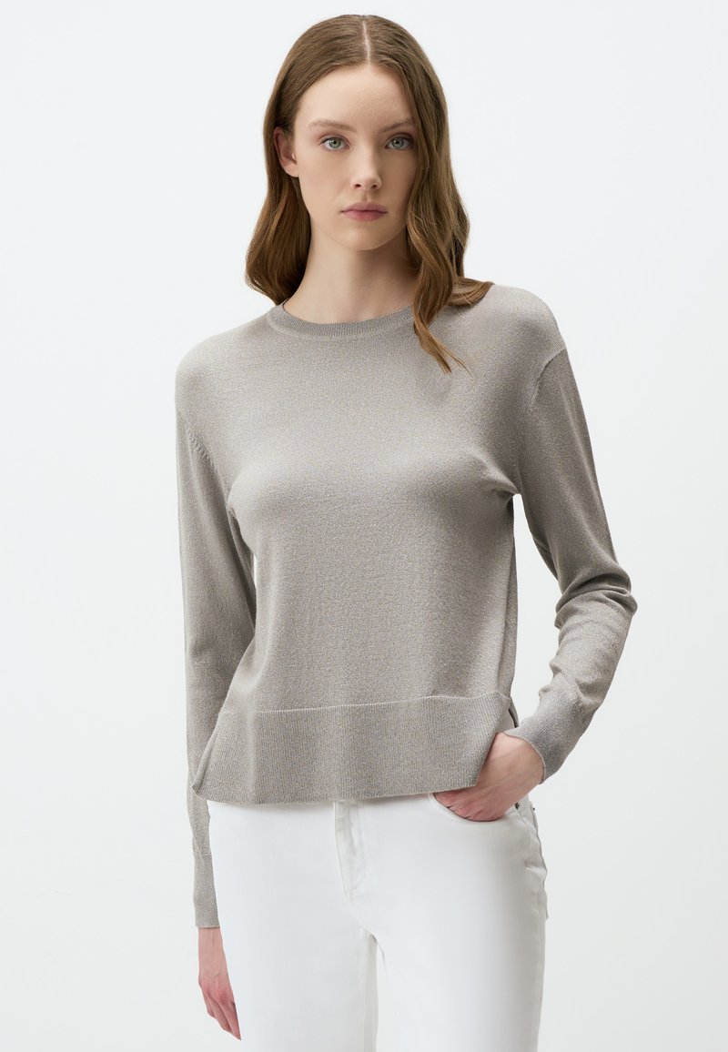 Pull long à manches longues gris clair avec un col rond et un tissu texturé. Il présente une coupe décontractée et un ourlet côtelé subtil, associé à un pantalon blanc.