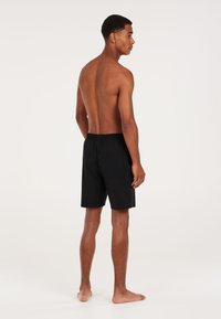 Zwarte zwemshorts met een elastische tailleband, knielange snit en gladde stof, gedragen door een persoon gezien van achteren.