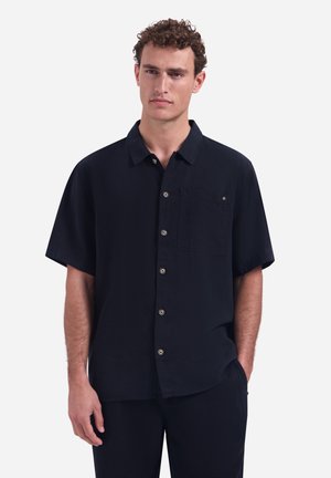 Junger Mann mit lockigem Haar, der ein schwarzes kurzärmeliges Hemd mit Brusttasche trägt, steht vor einem einfachen weißen Hintergrund.