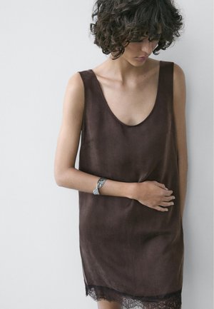 Frau mit lockigem Haar, die ein ärmelloses braunes Kleid mit Spitzenbesatz trägt, nach unten blickt und ihre Hand an der Taille auf einem einfarbigen Hintergrund ruhen lässt.