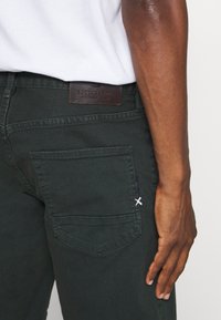 Ciemnozielone bawełniane spodnie cargo z brązową skórzaną naszywką w talii i ozdobnym przeszyciem na tylniej kieszeni.