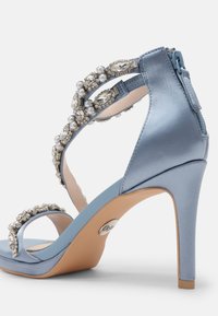 Sandale à talon haut en satin bleu clair ornée de bijoux décoratifs et de perles le long des sangles, avec une ouverture au niveau des orteils et une fermeture délicate à la cheville.