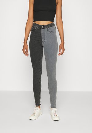 Frau trägt hoch taillierte Skinny-Jeans, ein Bein dunkelgrau und das andere hellgrau, kombiniert mit weißen Sneakers und einem schwarzen Crop-Top.