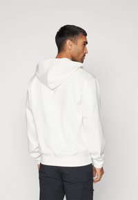 Haut à capuche blanc avec une poche kangourou, capuche réglable par cordon de serrage et poignets côtelés. Présente un petit logo à l'arrière. Texture douce.
