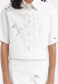 Biała krótka koszula dżinsowa z krótkimi rękawami, z ozdobnymi wycięciami i kieszonką z przodu. Klasyczne zapięcie na guziki.