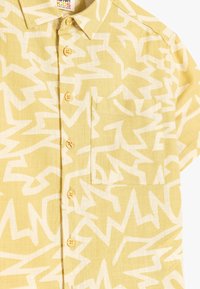Camisa de manga corta hecha de tejido ligero y texturizado en color amarillo pálido con un patrón de zigzag blanco; cuenta con un bolsillo en el frente y cierre de botones.