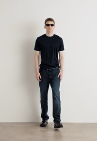 T-shirt nera a maniche corte, jeans scuri e occhiali da sole neri. Il modello è in piedi contro un muro grigio chiaro con uno sfondo minimalista.