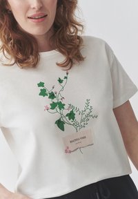 Bílý tričko s krátkým rukávem s designem zelené liány a růžovými květy a béžovou etiketou s nápisem "KRÁSNÉ LIÁNY." Měkký bavlněný materiál.