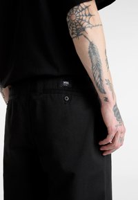 Schwarze Hosen mit einer hinteren Tasche und einem Knopf sowie einem Vans-Label. Haut mit sichtbaren Tätowierungen an einem Arm wird teilweise gezeigt.