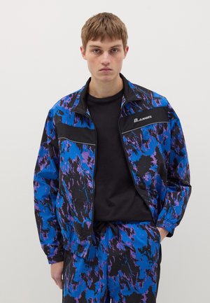 Trainingsjack gemaakt van lichtgewicht stof met een blauw en zwart abstract patroon, voorzien van een rits en zwarte accenten. Gekocht met bijpassende broek.
