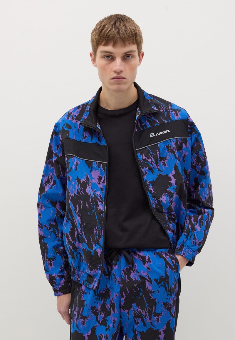 Giacca da pista realizzata in tessuto leggero con un motivo astratto blu e nero, caratterizzata da una chiusura a zip e dettagli neri. Abbinata a pantaloni coordinati.