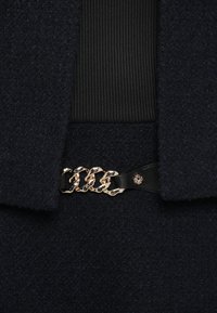 Donker gestructureerde wollen jas met een zwart geribd stoffen paneel en een zwarte leren riem met metalen schakels en een decoratief hardwaredetail.